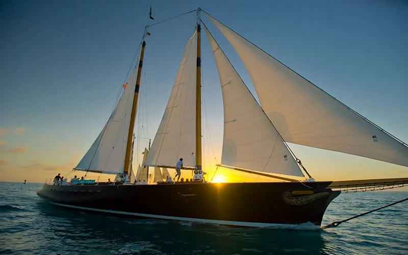 KeyWest_Champagne-Sunset_Slider-3 Schooner America 2.0 sailing with crew and guests on deck as the golden sun peaks through the sails for a Key West Sunset Sailing experience
