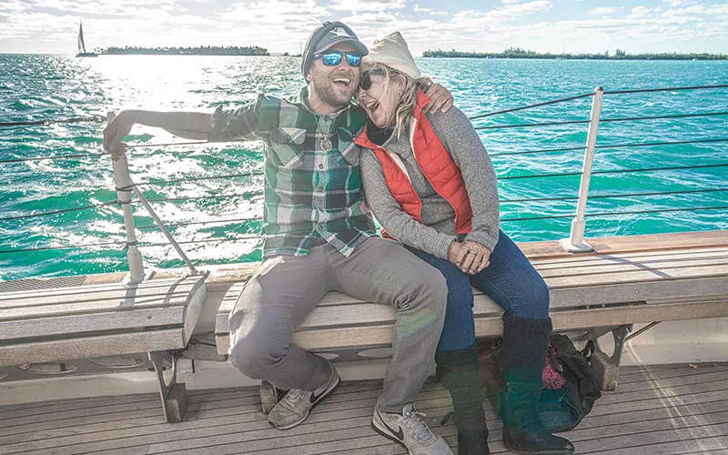 KeyWest_Champagne-Sunset_Slider-7 a couple hugging and laughing while sitting on a bench aboard the schooner america 2.0
