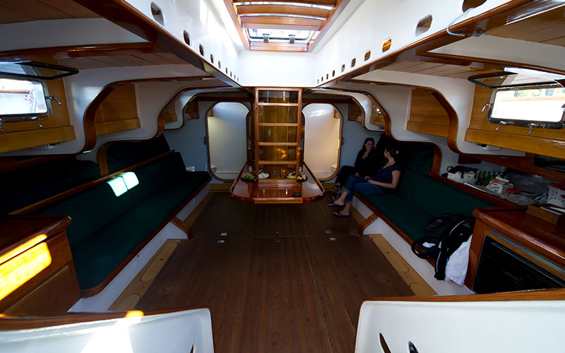 a lower deck view of the schooner america 2.0, with stairs to the upper deck as well as a small kitchen, table, and several benches